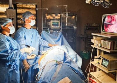 Surgeons in blue gowns and masks perform an operation in a dimly lit OR, monitors and medical equipment nearby.
