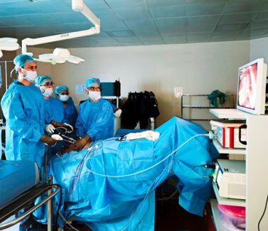 Surgical team in blue scrubs and masks performing an operation around a patient on the table; monitor displays the internal view being worked on.