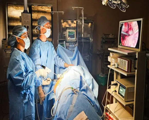 Two surgeons in blue gowns and masks perform surgery in an operating room, monitoring a patient with a screen showing an internal view of the area.